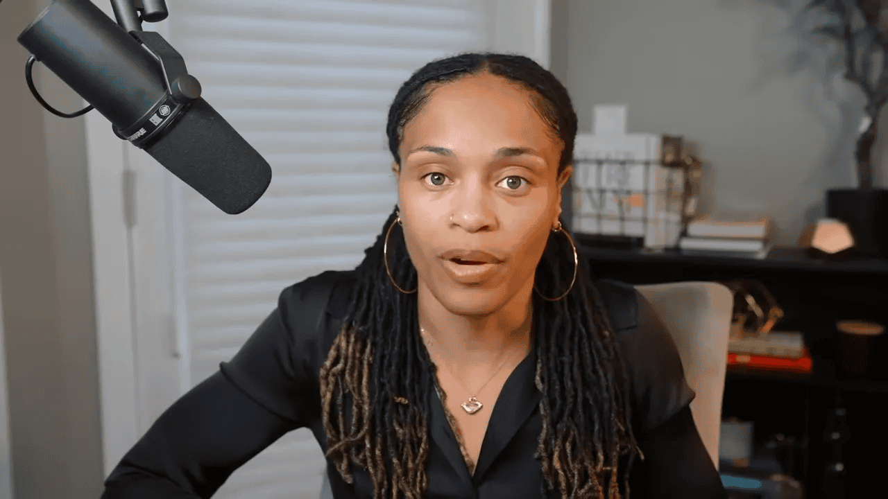 Presenter looking directly at camera with neutral, composed expression and microphone visible