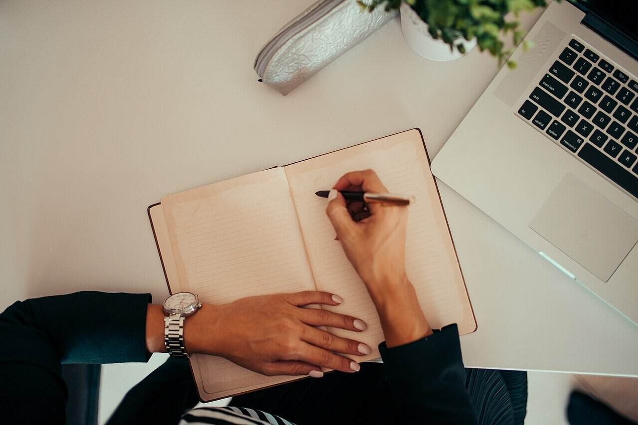 Person writing in a notebook beside an open laptop at a modern desk