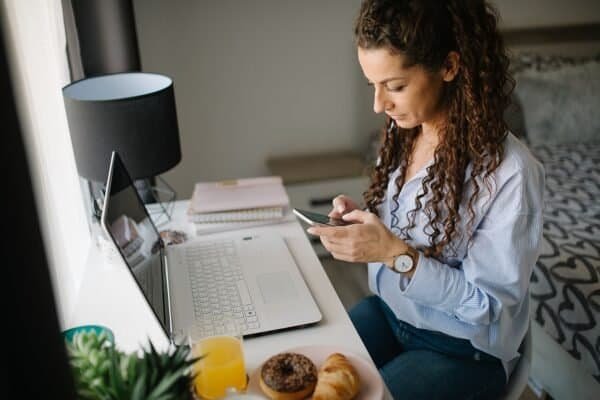 Woman using a smartphone at a home desk with a laptop and breakfast nearby